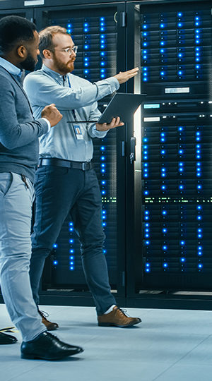 Men discussing something in a server room while holding a laptop.