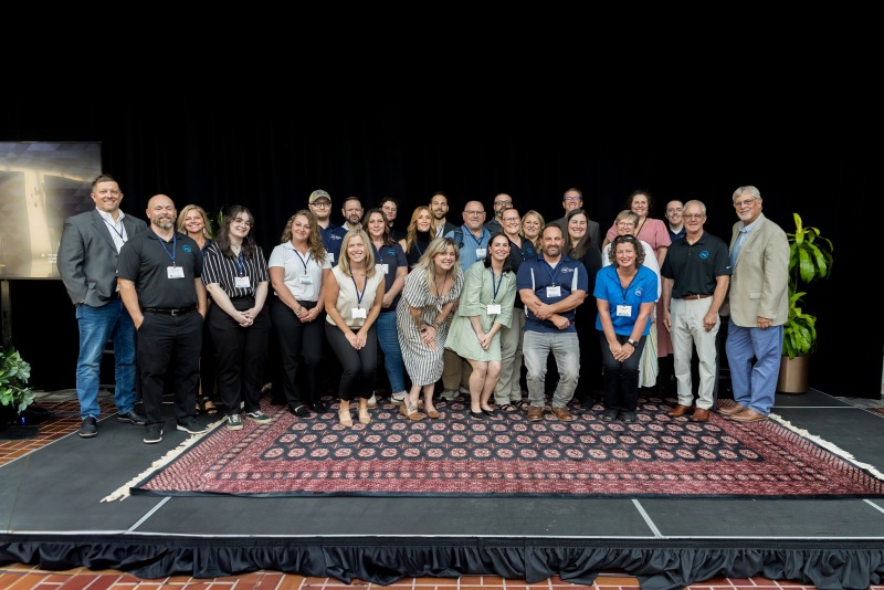 large group of adults poses together on a stage in front of a black backdrop. They are standing and kneeling in two rows, smiling at the camera. Most are wearing business casual attire with conference name badges. The stage is covered with a patterned rug, and there are potted plants on either side. The setting appears to be a professional event or conference gathering.