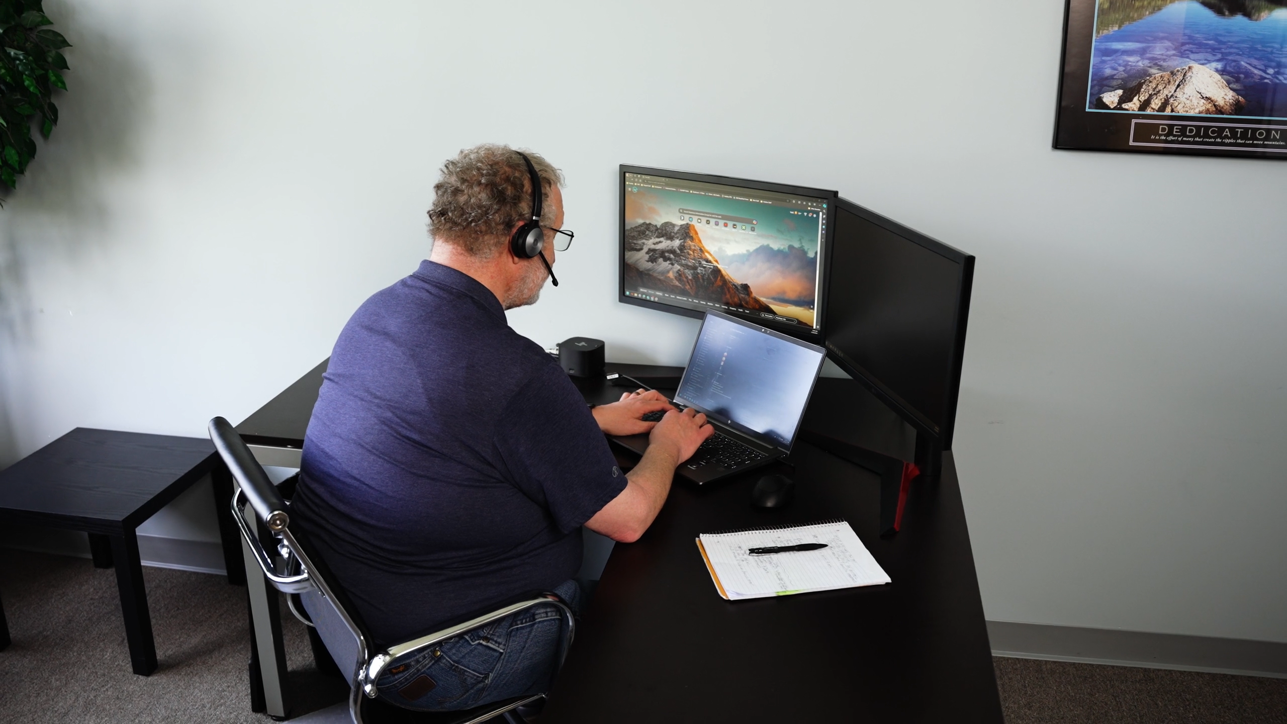 Man sitting at a desk in an office looking at three computer screens with a headset on.