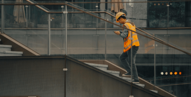 Construction worker in orange safety vest and hard hat using smartphone on job site stairs at modern building