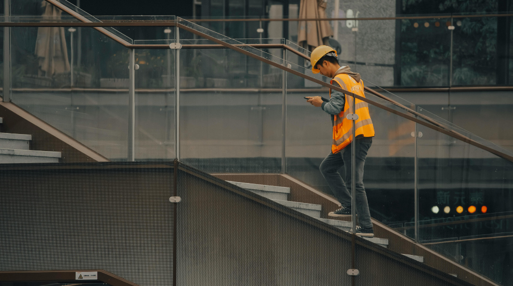 Construction worker in orange safety vest and hard hat using smartphone on job site stairs at modern building