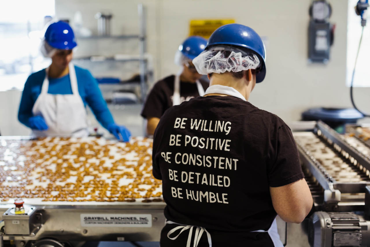 Food processing workers wearing blue hair nets and safety equipment work at a production line with baked goods. The worker in focus wears a black t-shirt with white text reading 'BE WILLING BE POSITIVE BE CONSISTENT BE DETAILED BE HUMBLE' while standing at an industrial conveyor belt in a commercial kitchen facility.