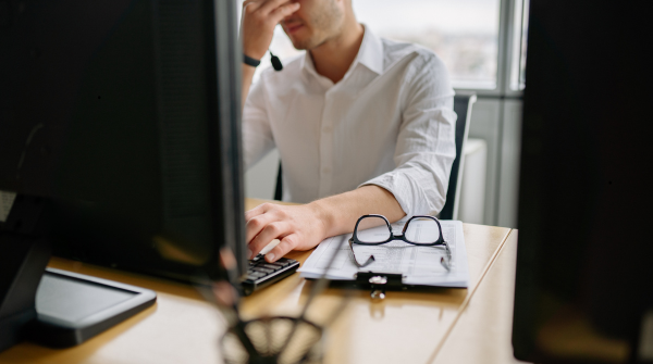 Business professional in a white dress shirt sitting at a desk working on a laptop with hand on forehead in a gesture suggesting stress or concentration. Reading glasses and paperwork rest on the wooden desk surface, with a computer monitor visible on the left side. The image conveys workplace challenges or problem-solving.
