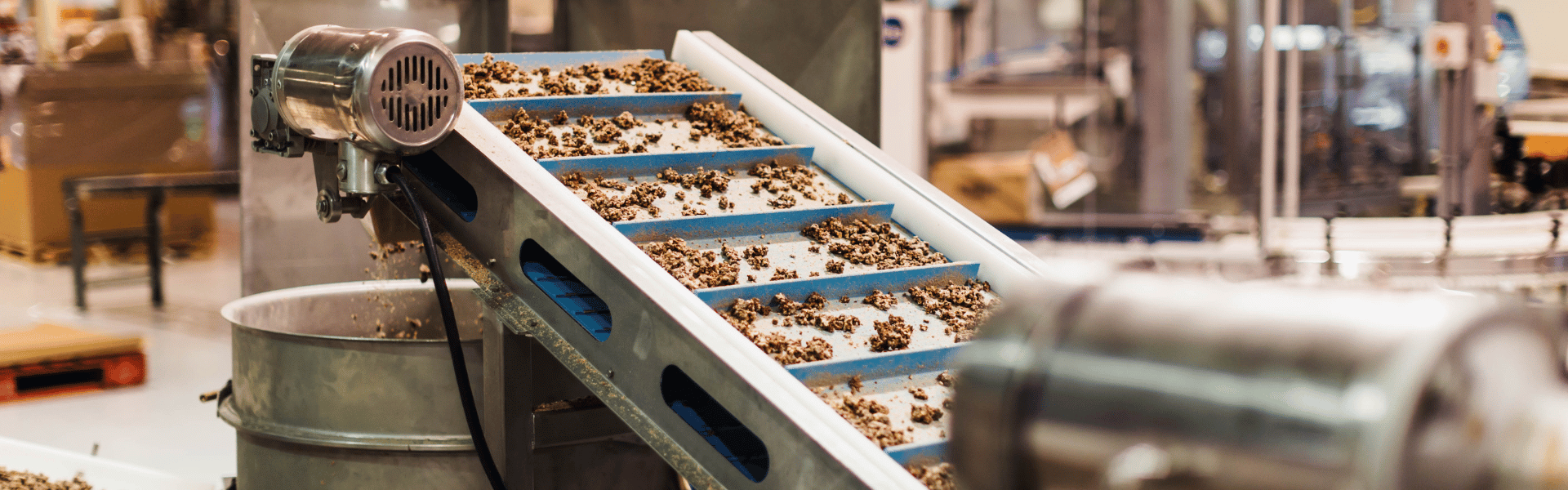 ndustrial vibrating conveyor system transporting chocolate or granola clusters up a multi-tiered inclined ramp with blue-lined channels in a commercial food production facility. The stainless steel equipment features a circular motor housing and collection basin, with additional processing machinery visible in the background of the manufacturing floor.