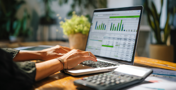 Close-up of hands typing on a laptop displaying business analytics dashboard with green bar charts and spreadsheet data. A calculator and financial documents are visible on the wooden desk alongside a potted plant in a bright office environment.