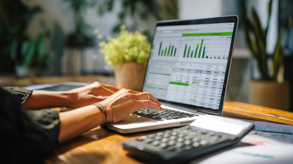 Close-up of hands typing on a laptop displaying business analytics dashboard with green bar charts and spreadsheet data. A calculator and financial documents are visible on the wooden desk alongside a potted plant in a bright office environment.