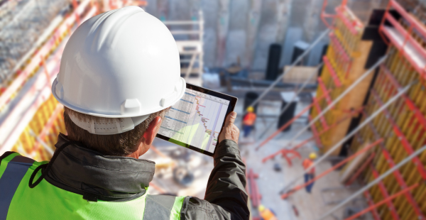 Construction worker in white hard hat and high-visibility safety vest views building plans on a tablet while overlooking an active construction site from above. The site shows steel framework, safety barriers, and construction materials scattered across the work area.