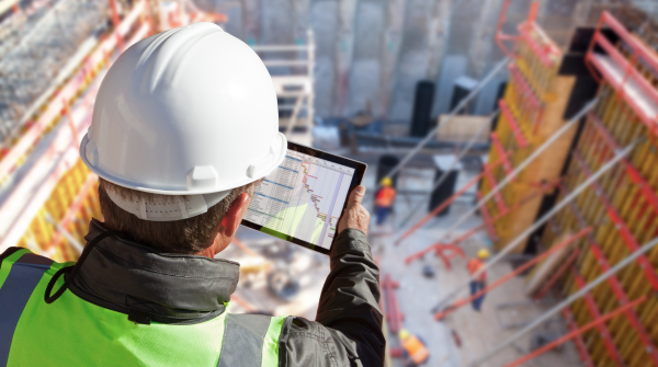 Construction worker in white hard hat and high-visibility safety vest views building plans on a tablet while overlooking an active construction site from above. The site shows steel framework, safety barriers, and construction materials scattered across the work area.