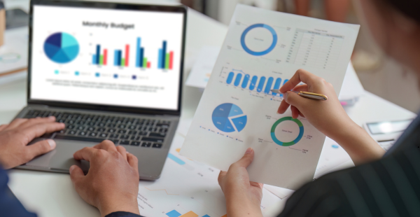 Two people collaborating over financial data at a desk. One person types on a laptop displaying a monthly budget dashboard with pie charts and bar graphs, while another reviews printed analytics reports featuring blue pie charts and bar graphs, holding a pen to annotate the documents.