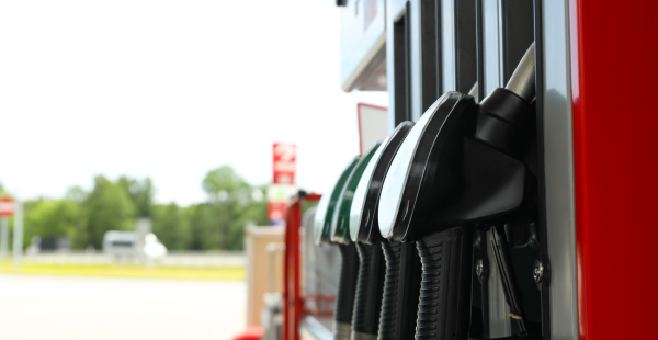 Close-up of multiple black fuel pump nozzles hanging at a gas station. The pumps are mounted on a red and silver dispenser, with an open field and trees visible in the blurred background under bright daylight.