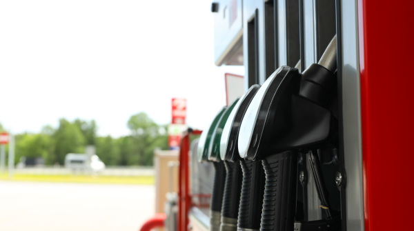 Close-up of multiple black fuel pump nozzles hanging at a gas station. The pumps are mounted on a red and silver dispenser, with an open field and trees visible in the blurred background under bright daylight.