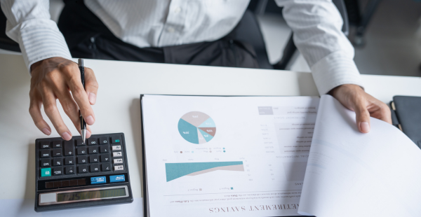 Overhead view of a businessperson in white dress shirt working at a desk, using a calculator with one hand while reviewing a financial report with the other. The report displays pie charts and bar graphs with blue and teal data visualizations on white paper.