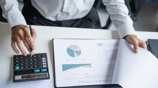 Overhead view of a businessperson in white dress shirt working at a desk, using a calculator with one hand while reviewing a financial report with the other. The report displays pie charts and bar graphs with blue and teal data visualizations on white paper.