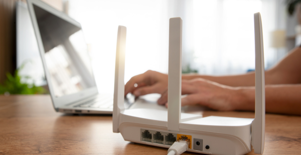 White wireless router with four antennas positioned on a wooden desk in the foreground, with a person's hands typing on a laptop keyboard in the blurred background. The router's ethernet ports and indicator lights are visible on its back panel.