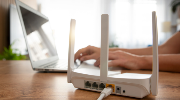 White wireless router with four antennas positioned on a wooden desk in the foreground, with a person's hands typing on a laptop keyboard in the blurred background. The router's ethernet ports and indicator lights are visible on its back panel.