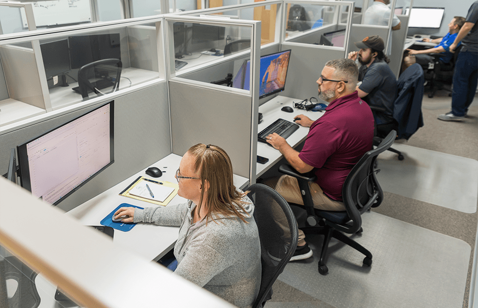 Office employees working at individual computer workstations separated by glass partitions. A woman wearing glasses works at a desk in the foreground while other team members focus on their monitors in the background.