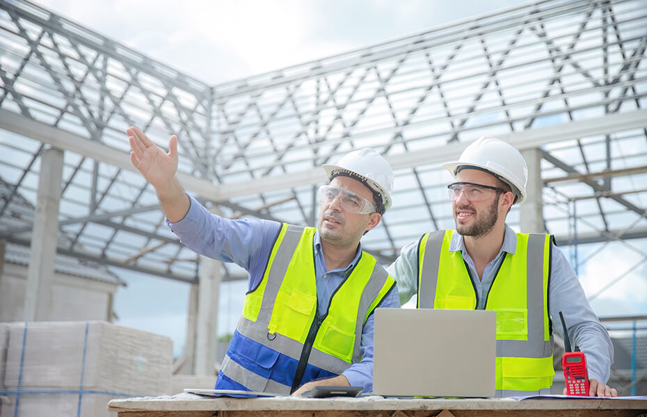Two construction workers wearing white hard hats, safety glasses, and yellow reflective safety vests stand on a building site reviewing a laptop. One worker points upward toward exposed steel beam framework with skylights overhead, while the other holds the computer. Building plans are visible on the table in front of them at the industrial construction site.