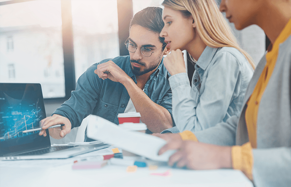 Small team of young professionals collaborating at a desk while viewing a laptop screen. They wear business casual clothing and appear engaged in creative or strategic work together in an office environment.