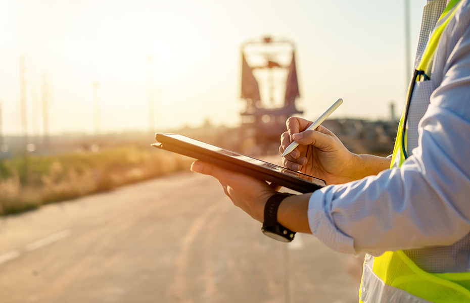 Construction worker wearing a white hard hat, yellow reflective safety vest, and light blue work shirt holding an iPad at a job site. A heavy crane is visible in the blurred background during golden hour sunset lighting.