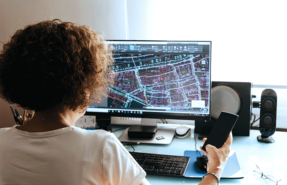 Person with curly hair wearing a white t-shirt and headphones sits at a desk holding a smartphone while viewing a desktop monitor displaying a detailed geographic information system (GIS) map showing urban streets, buildings, and infrastructure. The workspace includes a keyboard, computer speakers, mouse, and documents, illustrating digital mapping technology and mobile workflow integration.