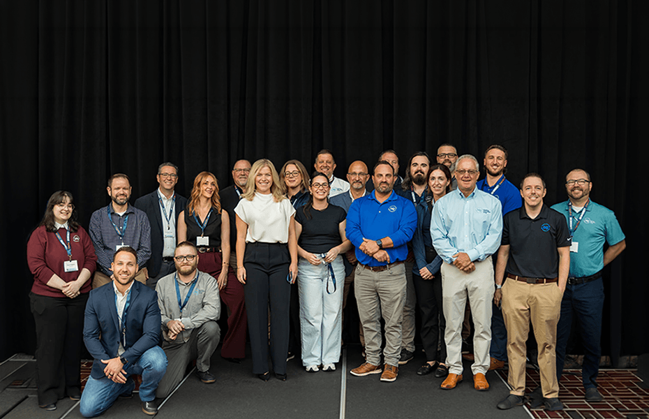 Diverse group of approximately 20 professionals standing together for a team photo at BIG university event, wearing business casual and formal attire with identification badges on lanyards, posed in front of a black curtain backdrop.